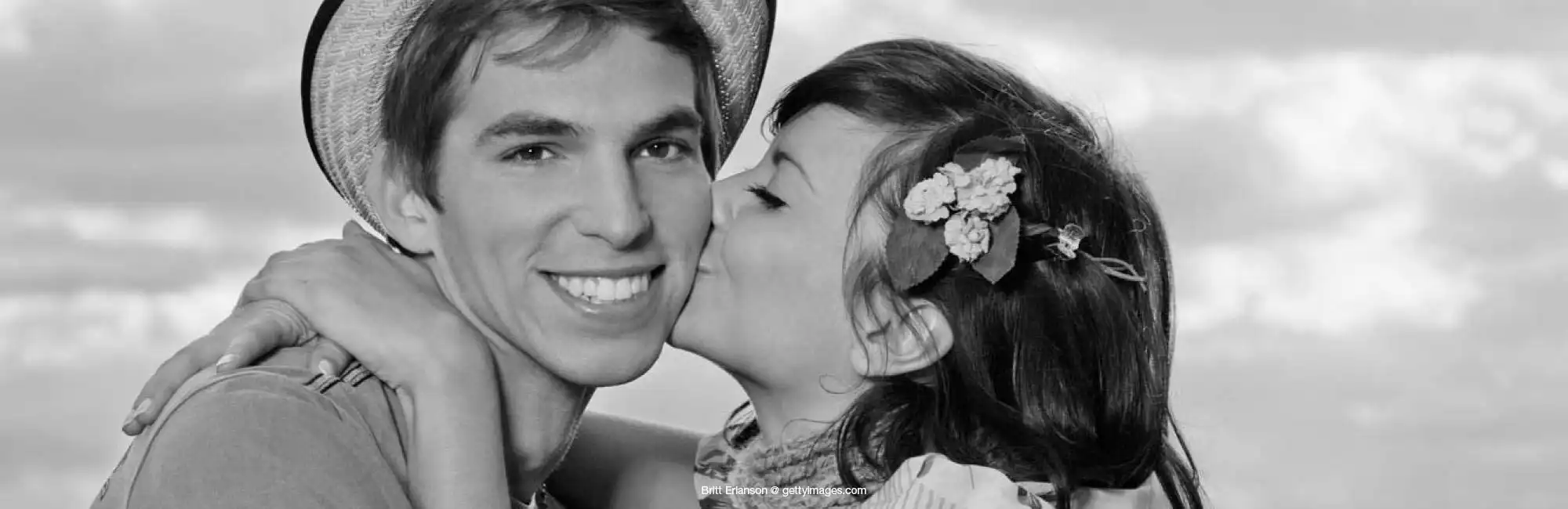 Über uns - Schwarz-Weiß-Vintage-Foto eines jungen Paars. Er trägt einen Strohhut und lächelt in die Kamera, sie trägt Blumen im Haar, umarnt und küsst ihn auf die Wange.